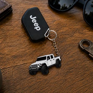 Op de afbeelding: Een zwarte Jeep-sleutelhanger met het woord "Jeep" in witte letters. Een zilveren ketting verbindt de sleutelhanger met een witte Jeep-truckbedel met zwarte wielen. Zonnebril en portemonnee zijn ook zichtbaar.