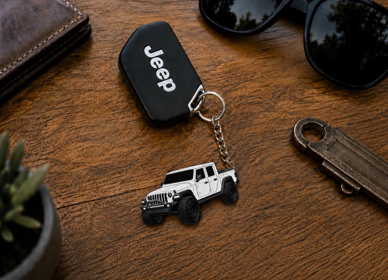 Op de afbeelding: Een zwarte Jeep-sleutelhanger met het woord "Jeep" in witte letters. Een zilveren ketting verbindt de sleutelhanger met een witte Jeep-truckbedel met zwarte wielen. Zonnebril en portemonnee zijn ook zichtbaar.