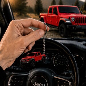 Op de afbeelding: Rode Jeep truck sleutelhanger met een zilveren ketting, gehouden voor het stuurwiel van een auto. Op de achtergrond staat een rode Jeep truck geparkeerd. Het stuurwiel heeft het Jeep-logo.