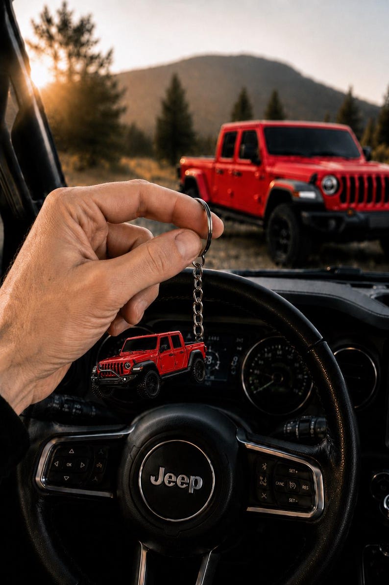 Op de afbeelding: Rode Jeep truck sleutelhanger met een zilveren ketting, gehouden voor het stuurwiel van een auto. Op de achtergrond staat een rode Jeep truck geparkeerd. Het stuurwiel heeft het Jeep-logo.