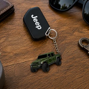 Op de afbeelding: Een zwarte Jeep-sleutelhanger met het woord 'Jeep' in witte letters. Een miniatuur groene Jeep-truck sleutelhanger hangt aan de sleutelhanger. Zonnebril, portemonnee en een metalen flesopener liggen ook op het houten oppervlak.