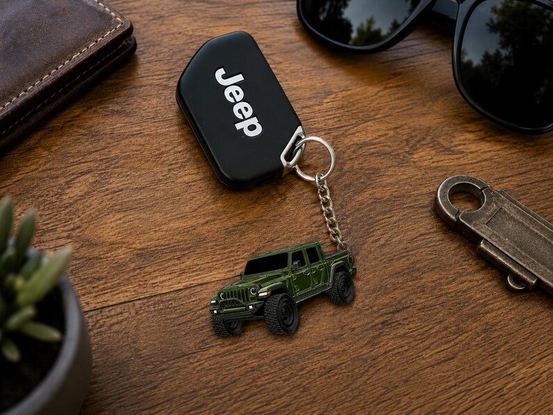 Op de afbeelding: Een zwarte Jeep-sleutelhanger met het woord 'Jeep' in witte letters. Een miniatuur groene Jeep-truck sleutelhanger hangt aan de sleutelhanger. Zonnebril, portemonnee en een metalen flesopener liggen ook op het houten oppervlak.