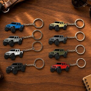 Op de afbeelding: Een verzameling miniatuur Jeep truck sleutelhangers in verschillende kleuren, waaronder blauw, geel, grijs, wit, groen, zwart en rood. Elke sleutelhanger heeft een gedetailleerd truckontwerp met een zilveren sleutelring en ketting. Gerangschikt op een houten oppervlak.