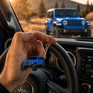 May include: A miniature blue off-road vehicle keychain featuring a detailed body, black wheels, and a silver metal chain. The keychain is held in front of a black steering wheel with a Jeep logo, with a full-sized blue Jeep visible in the background.