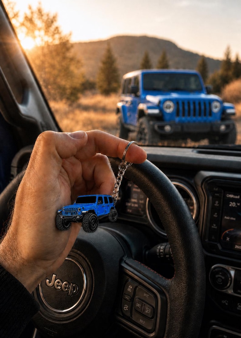 May include: A miniature blue off-road vehicle keychain featuring a detailed body, black wheels, and a silver metal chain. The keychain is held in front of a black steering wheel with a Jeep logo, with a full-sized blue Jeep visible in the background.