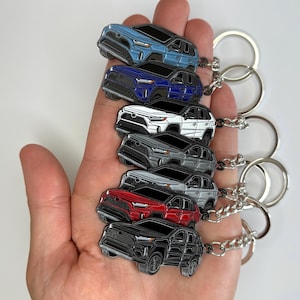 Puede incluir: Una colección de llaveros con forma de coche en varios colores, incluyendo azul, marino, blanco, gris, rojo y negro. Cada llavero presenta un diseño detallado de coche con un llavero plateado adjunto. Los llaveros se sostienen en una mano sobre un fondo blanco.