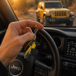 May include: A yellow metal keychain shaped like a four-door off-road vehicle with black wheels and detailing, attached to a silver-toned metal split ring and chain, held against the backdrop of a steering wheel and a matching yellow vehicle.