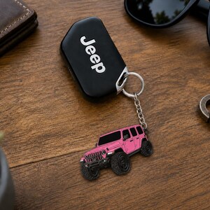 May include: A black Jeep key fob attached to a silver metal chain and a pink off-road vehicle charm. The charm features a detailed pink SUV with black wheels, a front grille, and windows. The key fob displays the white Jeep logo on its surface.