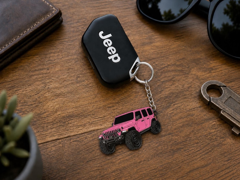 May include: A black Jeep key fob attached to a silver metal chain and a pink off-road vehicle charm. The charm features a detailed pink SUV with black wheels, a front grille, and windows. The key fob displays the white Jeep logo on its surface.