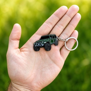 May include: A dark green metal keychain shaped like a four-door off-road vehicle with black tires, a front grille, and a silver-toned circular split ring attached by a short metal chain, showcasing a detailed miniature automotive design.