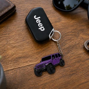 May include: A black Jeep key fob with white text attached to a silver metal chain and a purple SUV-shaped charm. The charm features a detailed purple vehicle with black wheels, a front grille, and windows, connected by a small circular ring.