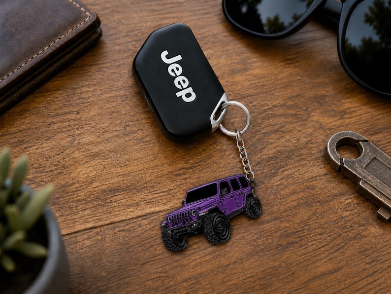 May include: A black Jeep key fob with white text attached to a silver metal chain and a purple SUV-shaped charm. The charm features a detailed purple vehicle with black wheels, a front grille, and windows, connected by a small circular ring.