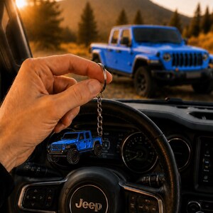 Op de afbeelding: Een blauwe Jeep-trucksleutelhanger wordt voor een blauwe Jeep-truck gehouden. De sleutelhanger is een miniatuurversie van de truck, met zwarte banden en een zilveren ketting. De achtergrond toont een berglandschap en het Jeep-stuurwiel.