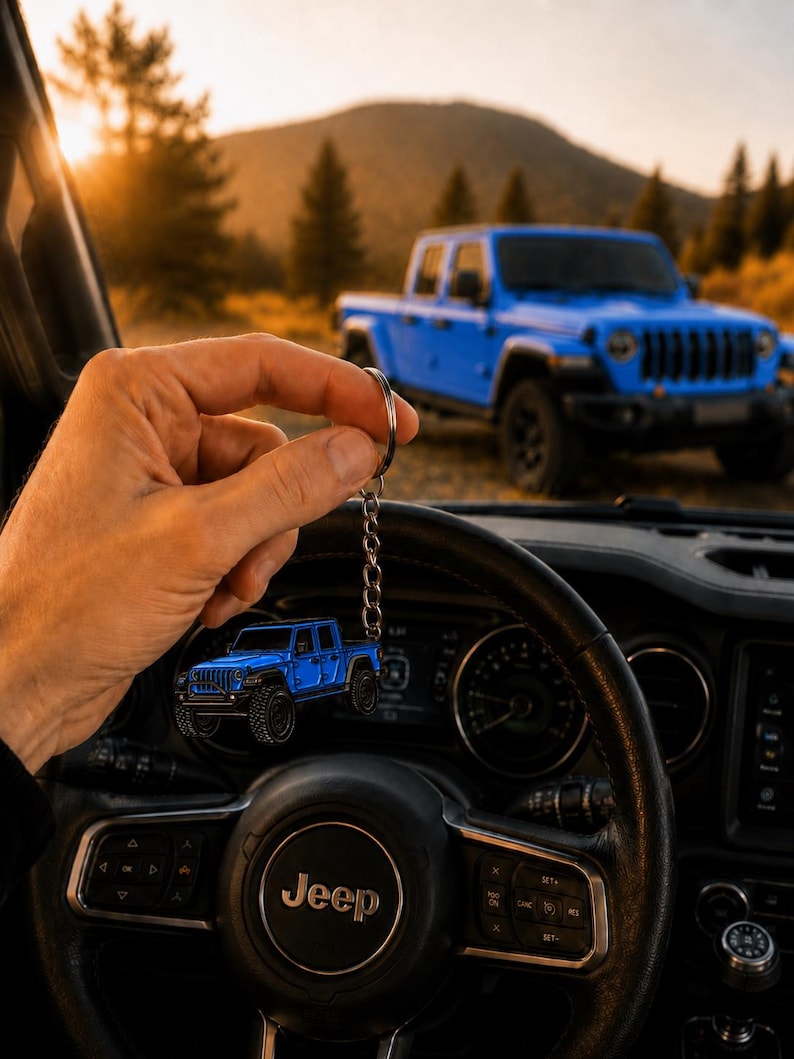 Op de afbeelding: Een blauwe Jeep-trucksleutelhanger wordt voor een blauwe Jeep-truck gehouden. De sleutelhanger is een miniatuurversie van de truck, met zwarte banden en een zilveren ketting. De achtergrond toont een berglandschap en het Jeep-stuurwiel.
