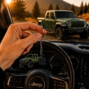 Op de afbeelding: Een groene Jeep-truck sleutelhanger wordt voor een Jeep-stuurwiel gehouden. De sleutelhanger weerspiegelt een groene Jeep-truck die op de achtergrond geparkeerd staat. Het stuurwiel heeft het Jeep-logo.
