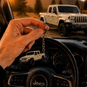 Op de afbeelding: Een sleutelhanger met een beige Jeep pick-up truck bedel. De bedel wordt voor een Jeep stuurwiel gehouden, met een bijpassende Jeep truck op de achtergrond. De sc&egrave;ne speelt zich buiten af met een zonsondergang.
