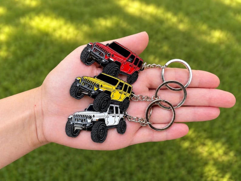 May include: Three Jeep keychains in red, yellow, and white, each with black wheels and a silver keyring. The keychains are held in a hand against a blurred green background.