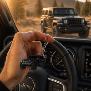 May include: A black metal keychain shaped like a Jeep Wrangler SUV, featuring detailed tires, a front grille, and headlights, attached to a silver-toned circular split ring, held up against the backdrop of a vehicle interior and a parked SUV.