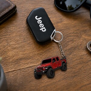 Op de afbeelding: Zwarte Jeep-sleutelhanger met een zilveren sleutelring en een rode Jeep-truck-sleutelhanger. De sleutelhanger heeft het woord "Jeep" in het wit. De vrachtwagen-sleutelhanger is een gedetailleerd miniatuurmodel.