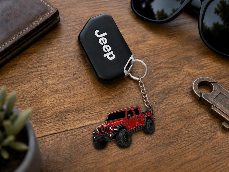 Op de afbeelding: Zwarte Jeep-sleutelhanger met een zilveren sleutelring en een rode Jeep-truck-sleutelhanger. De sleutelhanger heeft het woord "Jeep" in het wit. De vrachtwagen-sleutelhanger is een gedetailleerd miniatuurmodel.