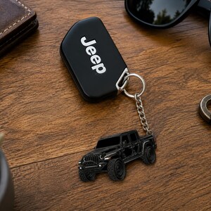 Op de afbeelding: Zwarte Jeep-sleutelhanger met bijpassende zwarte Jeep-trucksleutelhanger. De sleutelhanger heeft het woord "Jeep" in het wit. De sleutelhanger is bevestigd met een zilveren ketting en ring. De items liggen op een houten oppervlak.