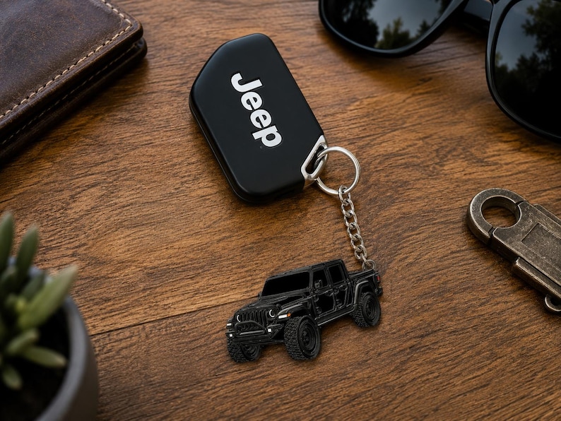 Op de afbeelding: Zwarte Jeep-sleutelhanger met bijpassende zwarte Jeep-trucksleutelhanger. De sleutelhanger heeft het woord "Jeep" in het wit. De sleutelhanger is bevestigd met een zilveren ketting en ring. De items liggen op een houten oppervlak.