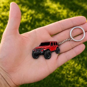 May include: A red off-road vehicle keychain featuring a detailed four-door SUV design with black wheels, a front grille, and a silver metal split ring attached by a short chain, showcasing a realistic miniature representation of a rugged automobile.