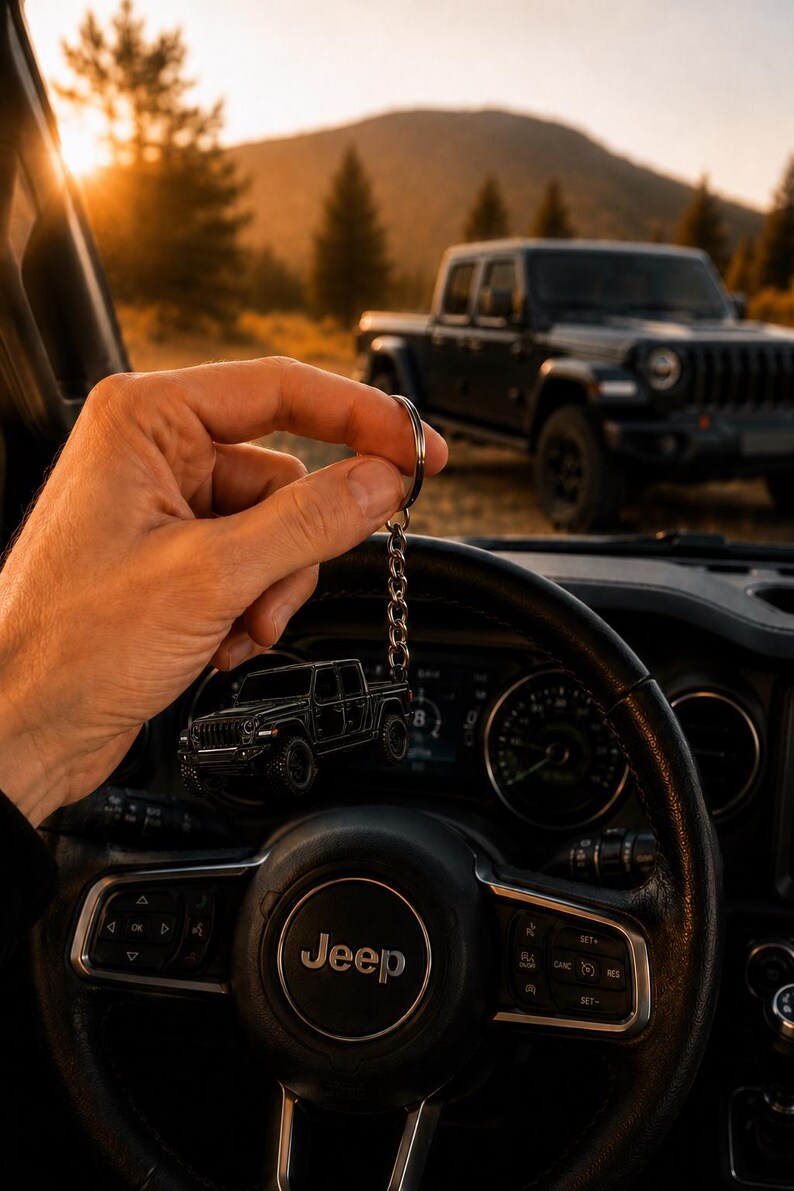 Op de afbeelding: Een zwarte Jeep truck sleutelhanger wordt voor een Jeep stuurwiel gehouden. De sleutelhanger is een miniatuur replica van de truck. De achtergrond toont een zwarte Jeep truck geparkeerd in de buitenlucht met een berg en zonsondergang.