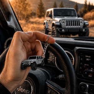 May include: A silver-colored metal keychain shaped like a four-door off-road vehicle with black wheels and a detailed front grille, featuring a circular split ring and a short chain, held against the backdrop of a vehicle interior and landscape.