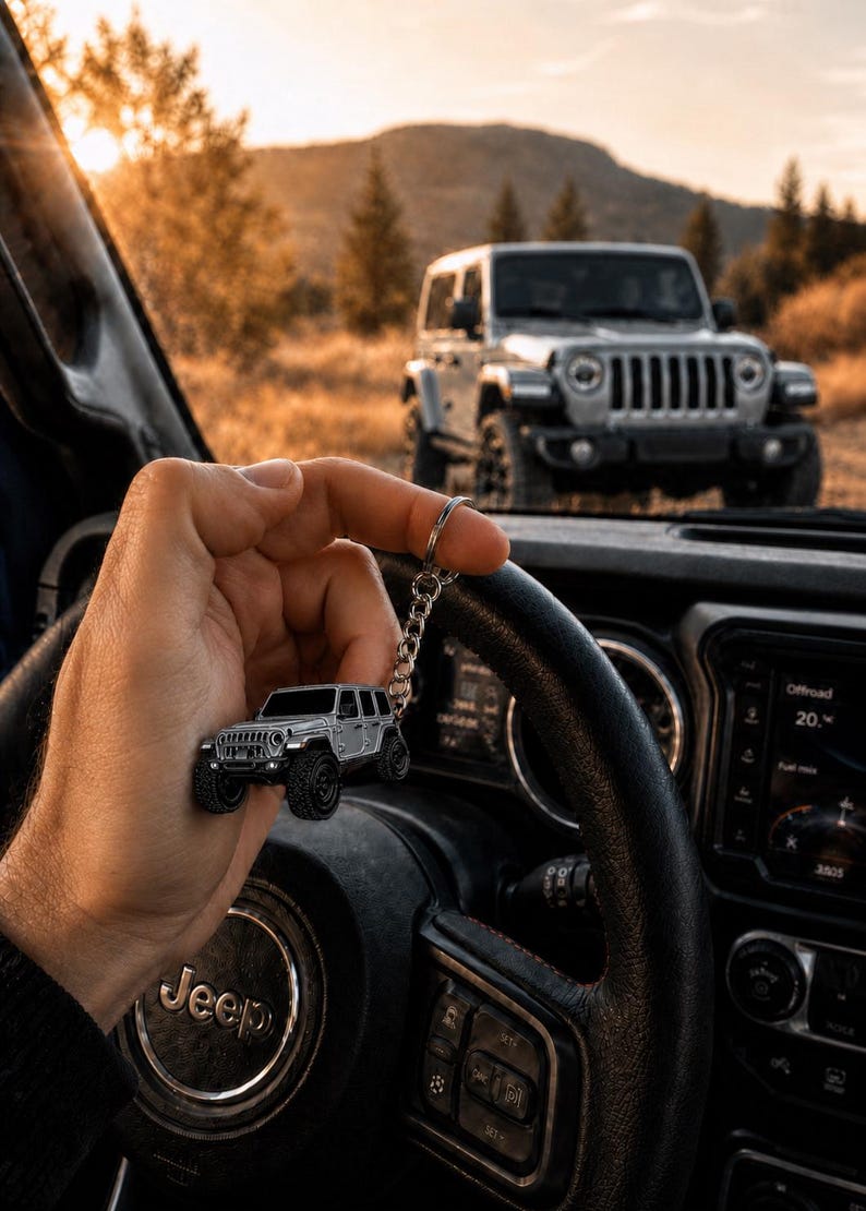 May include: A silver-colored metal keychain shaped like a four-door off-road vehicle with black wheels and a detailed front grille, featuring a circular split ring and a short chain, held against the backdrop of a vehicle interior and landscape.