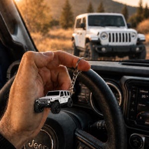 May include: A white off-road vehicle keychain featuring a detailed miniature model of a four-door SUV with black wheels and a silver metal chain ring, held up against the backdrop of a full-sized white SUV parked in a natural outdoor setting.
