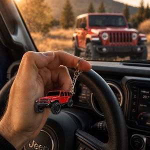 May include: A miniature red off-road vehicle keychain featuring a detailed metal body, black wheels, and a silver-toned metal chain and ring attachment. The keychain is held up against a blurred background showing a full-sized red vehicle.