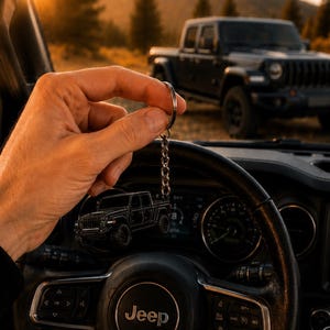 Op de afbeelding: Een zwarte Jeep truck sleutelhanger, gehouden voor een Jeep stuurwiel. De sleutelhanger is een gedetailleerde miniatuur van de truck. De achtergrond toont een zwarte Jeep truck geparkeerd in de buitenlucht, met een berg en zonsondergang in de verte.