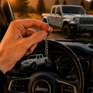 Op de afbeelding: Een zilveren Jeep truck sleutelhanger, gehouden voor het stuur van een auto. De sleutelhanger weerspiegelt een full-size truck die op de achtergrond geparkeerd staat. Het stuur is zwart met het Jeep-logo in het midden. De achtergrond toont een berglandschap.