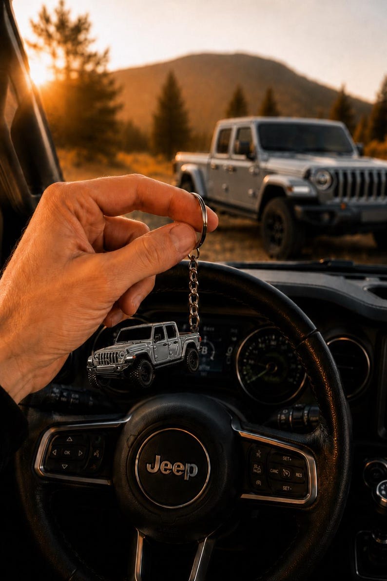 Op de afbeelding: Een zilveren Jeep truck sleutelhanger, gehouden voor het stuur van een auto. De sleutelhanger weerspiegelt een full-size truck die op de achtergrond geparkeerd staat. Het stuur is zwart met het Jeep-logo in het midden. De achtergrond toont een berglandschap.