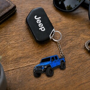 Op de afbeelding: Een zwarte Jeep-sleutelhanger met het woord 'Jeep' in witte letters. Een blauwe Jeep-trucksleutelhanger is bevestigd. De items liggen op een houten oppervlak met een zonnebril en een portemonnee.