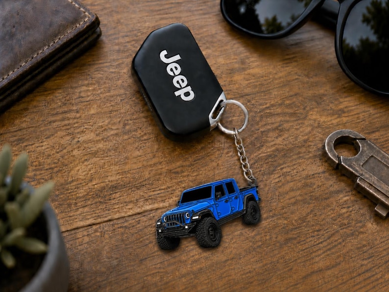 Op de afbeelding: Een zwarte Jeep-sleutelhanger met het woord 'Jeep' in witte letters. Een blauwe Jeep-trucksleutelhanger is bevestigd. De items liggen op een houten oppervlak met een zonnebril en een portemonnee.