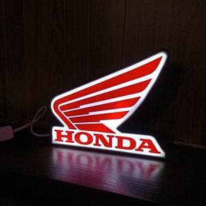 Op de afbeelding: Een verlicht bord met het Honda-logo in wit en rood. Het logo toont een gestileerde vleugel met drie lijnen en het woord "HONDA" in witte letters onder de vleugel.