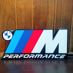 Może przedstawiać: Czarno-biały znak BMW Performance z logo BMW po lewej stronie. Znak posiada niebieskie, granatowe i czerwone paski, z białą literą "M" i słowem "PERFORMANCE" w kolorze białym.