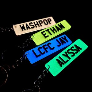 Op de afbeelding: Vier glow-in-the-dark sleutelhangers met verschillende namen: Washpop, Ethan, LCFC Jay en Alyssa. De sleutelhangers zijn bevestigd aan zilveren kettingen.