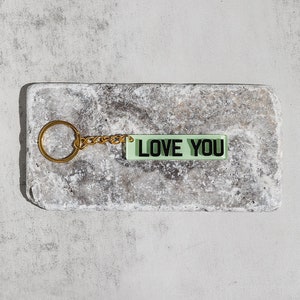 May include: A gold keychain with a green rectangular charm that says "LOVE YOU" in black letters.