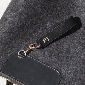 K&ouml;nnte beinhalten: Eine schwarze Filz-Tragetasche mit einem schwarzen Leder-Rei&szlig;verschluss-Zipper und einem schwarzen Leder-Patch mit einem goldenen Rei&szlig;verschluss. Die Tasche hat einen schwarzen Lederriemen mit einem goldenen Metallverschluss und ein schwarzes Leder-Etikett mit den Initialen "EJ" in goldenen Buchstaben.