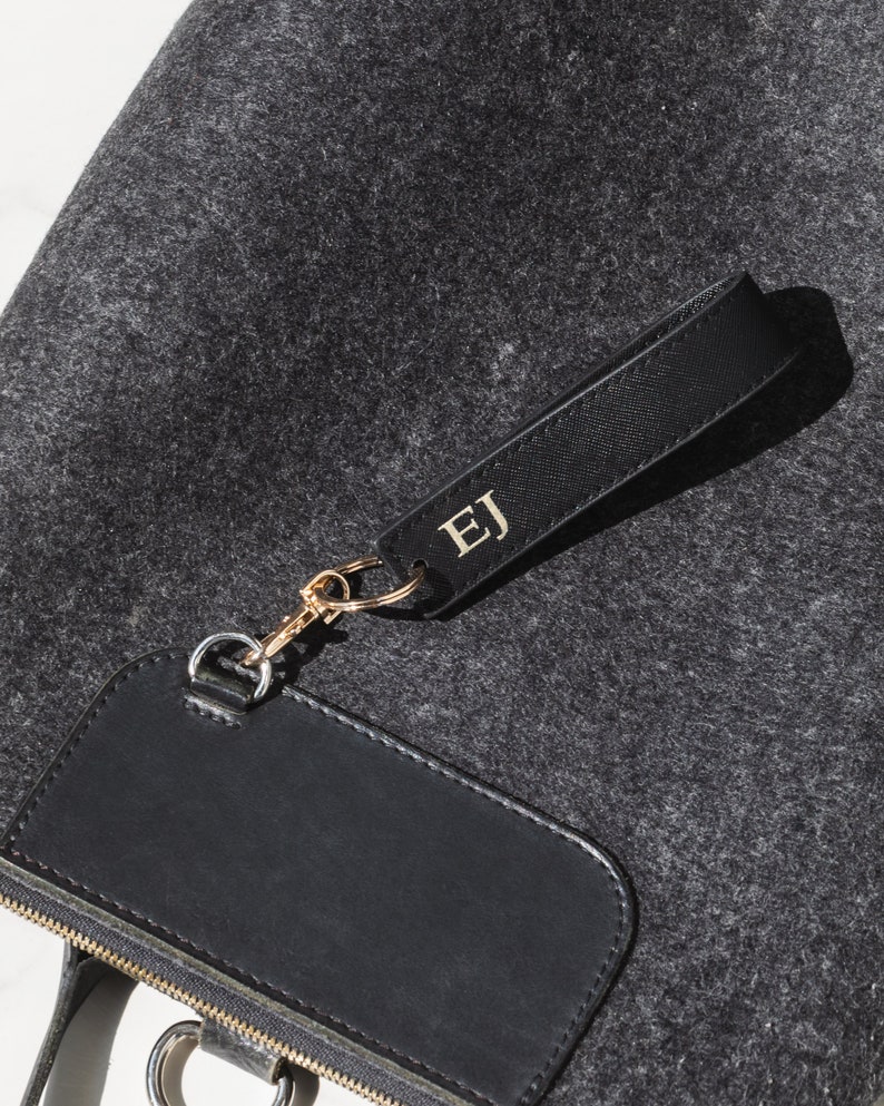 K&ouml;nnte beinhalten: Eine schwarze Filz-Tragetasche mit einem schwarzen Leder-Rei&szlig;verschluss-Zipper und einem schwarzen Leder-Patch mit einem goldenen Rei&szlig;verschluss. Die Tasche hat einen schwarzen Lederriemen mit einem goldenen Metallverschluss und ein schwarzes Leder-Etikett mit den Initialen "EJ" in goldenen Buchstaben.