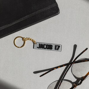May include: A gold keychain with a clear acrylic tag that says "JACOB 17" in black lettering. The keychain is lying on a gray surface next to a pair of tortoiseshell glasses.