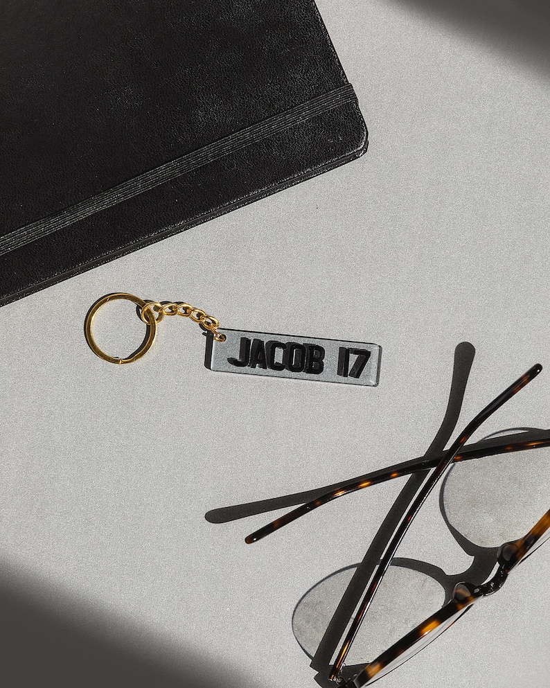 May include: A gold keychain with a clear acrylic tag that says "JACOB 17" in black lettering. The keychain is lying on a gray surface next to a pair of tortoiseshell glasses.
