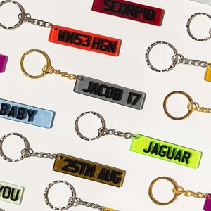 May include: A collection of colorful keychains with different text on each tag. The keychains are made of clear acrylic with black text. The text on the tags includes "ROSE", "SCORPIO", "WM53 HGN", "JACOB 17", "BABY", "25TH AUG", "JAGUAR", "KIGH XR", and "YOU".