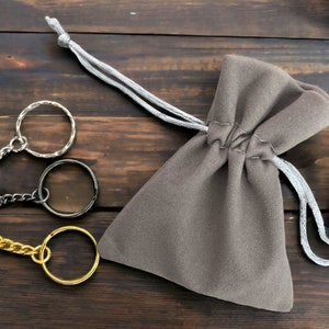 May include: Three silver keychains with different styles of rings and chains. The keychains are on a wooden surface and next to a gray drawstring bag.