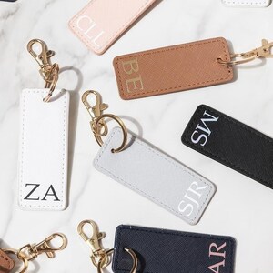 Op de afbeelding: Zes gepersonaliseerde leren sleutelhangers met gouden ringen en sluitingen. De sleutelhangers zijn in verschillende kleuren verkrijgbaar, waaronder wit, roze, bruin, grijs, zwart en marineblauw. Elke sleutelhanger heeft een andere set initialen in witte of roze letters.