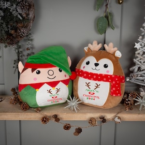 May include: Two plush Christmas decorations, one a green elf with a red hat and the other a brown reindeer with a red scarf. Both decorations have embroidered text that says "[Name]'s 1st Christmas 2023".