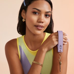 K&ouml;nnte beinhalten: Eine Frau tr&auml;gt ein gelbes und violettes, &auml;rmelloses Oberteil mit einem goldenen Gliederarmband. Sie h&auml;lt einen lilafarbenen Leder-Schl&uuml;sselanh&auml;nger mit den Initialen "CD" in der Hand.